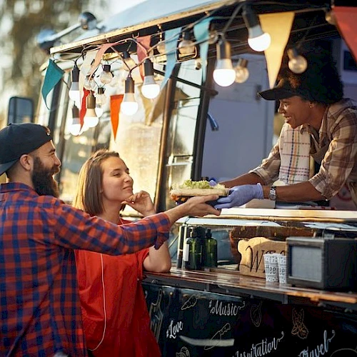 Two friends at a food truck hand off a wrapped burrito while the vendor serves: casual outdoor dining under string lights and bunting.