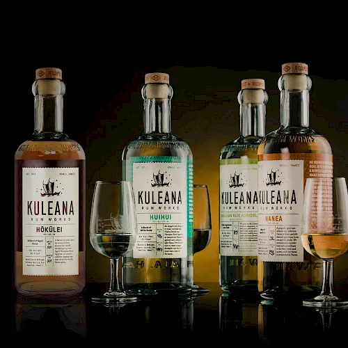 A row of Kuleana whiskey bottles with glasses, dark background, labels visible, three flavors.