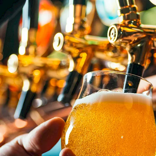 A hand holds a foamy beer in front of shiny beer taps, with warm golden lights in a bar.