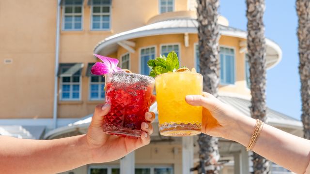 Two hands clink red and yellow iced cocktails, each topped with fruit garnish and mint, in bright outdoor sunshine by a building.