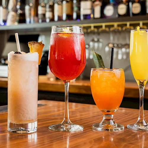 A row of colorful cocktails on a wooden bar, with bottles lined up behind the bar.