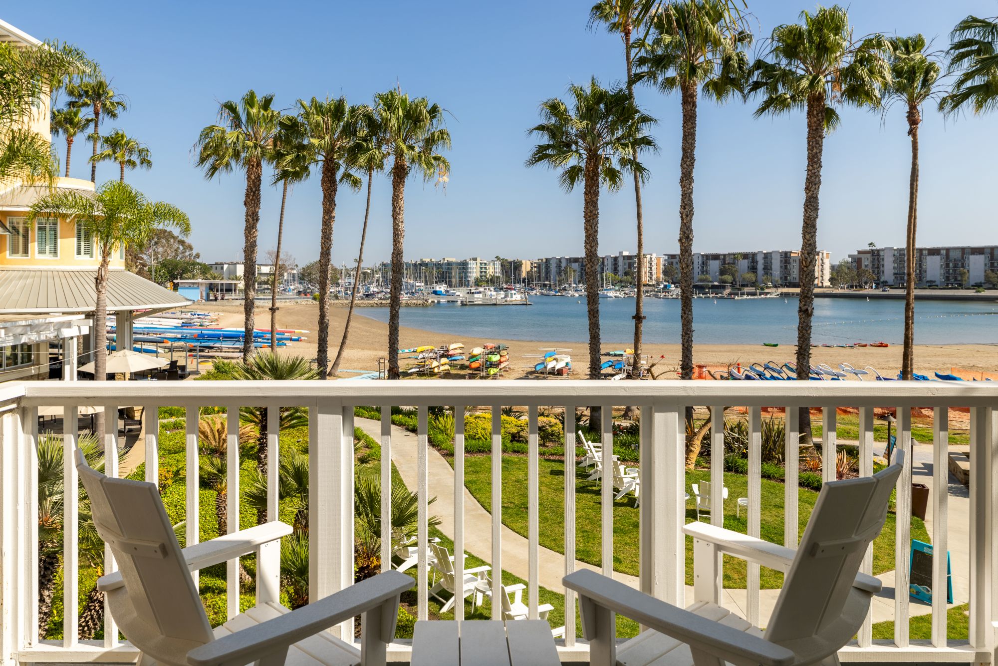 A sunny beach scene with tall palm trees, a sandy shore, and calm blue water viewed from a balcony with white chairs.