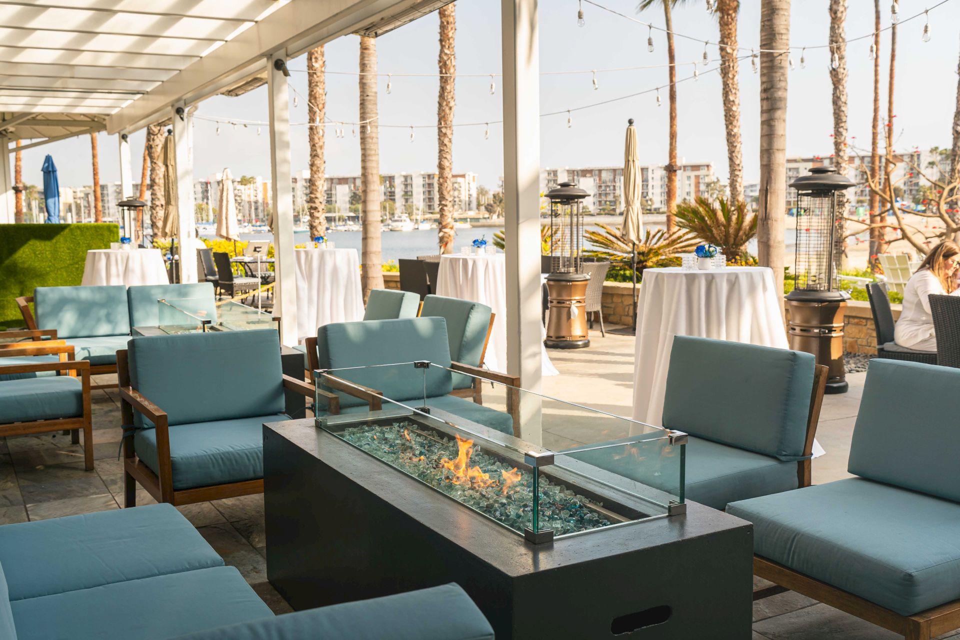 Outdoor patio lounge with blue cushions, a glass firepit with flames, tall palm trees, string lights, and heaters beside banquet tables.