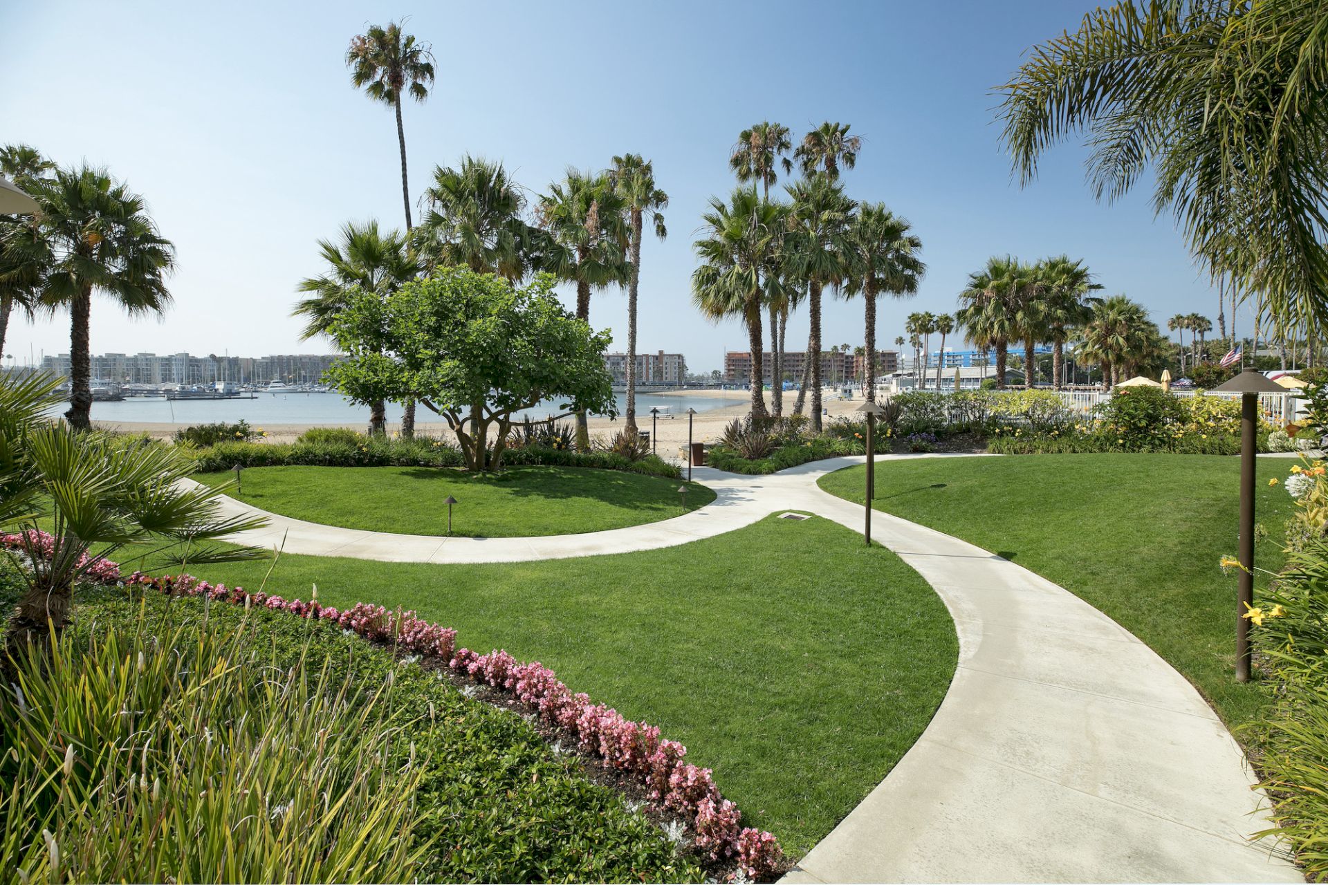 A sunny seaside garden with green lawns, winding walkways, palm trees, and sailboats along the bay.