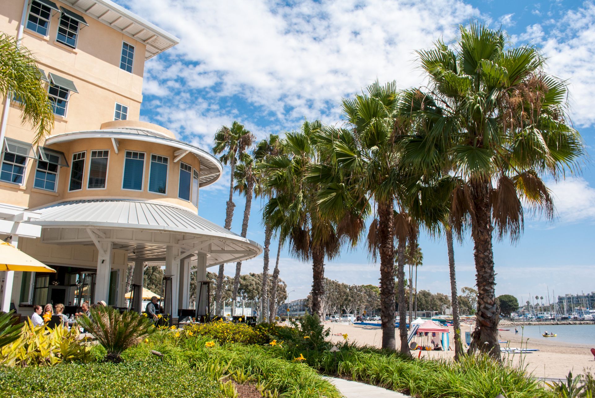 A sunny seaside hotel building with curved veranda, lush palm trees, and a marina with boats under a blue sky.