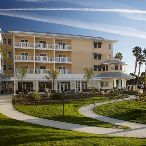 A sunny yellow hotel or resort with multiple balconies, palm trees, a curved walkway, and manicured lawns under a clear blue sky.