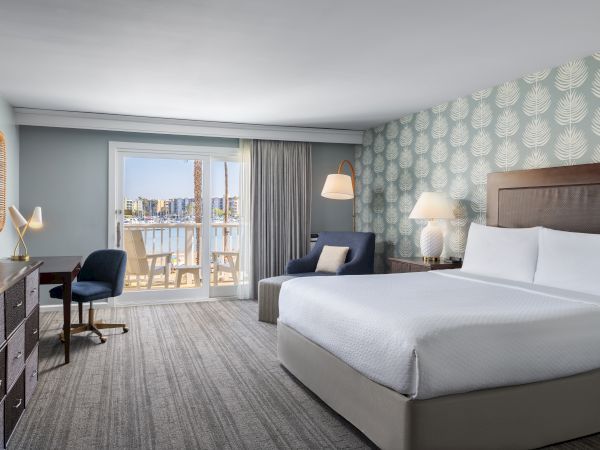 A modern hotel room with two queen beds, a desk, a seating area, and a balcony view of the city beyond soft, patterned wallpaper and neutral tones.