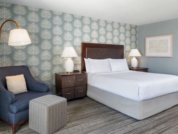A hotel room with a king bed, teal patterned accent wall, dark wood nightstands, twin lamps, a blue armchair, and a light gray ottoman.