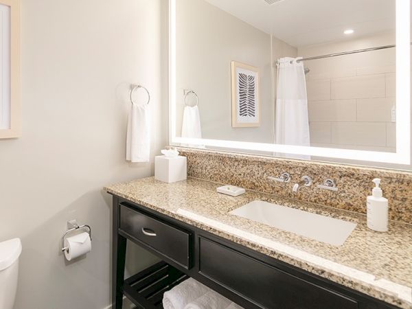 This image shows a modern bathroom with a granite countertop, sink, illuminated mirror, towel rack, and a shower curtain in the background.