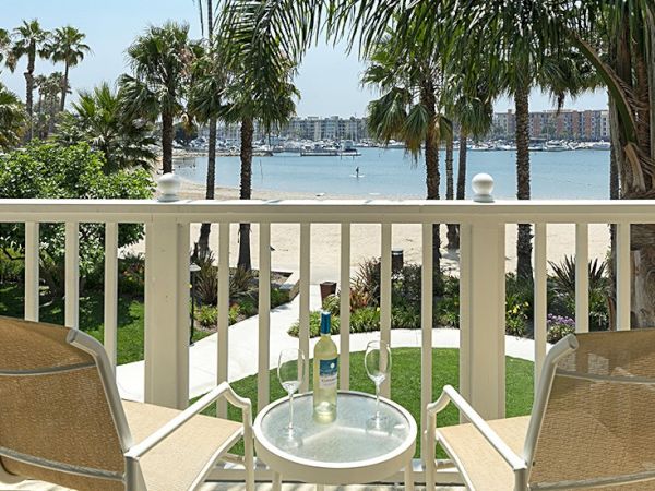 A balcony overlooking a beach with two chairs, a table, wine bottle, glasses, palm trees, and distant boats in the water.