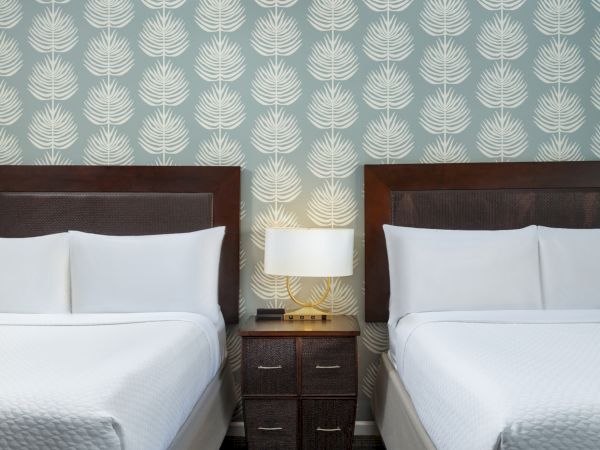 Two single beds with white linens, a shared nightstand, a lamp, and teal patterned wallpaper behind them, in a hotel room.
