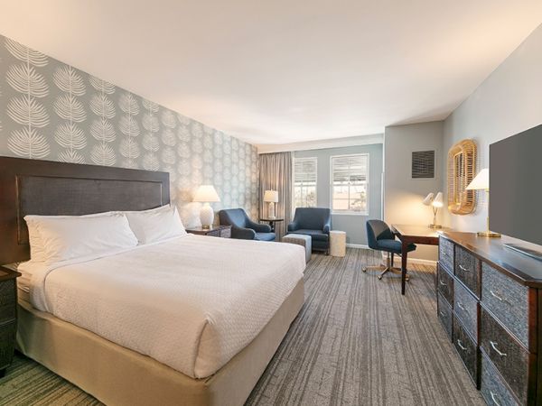 A hotel room with a bed, two chairs, a desk, and a TV, featuring patterned wallpaper and ample lighting.