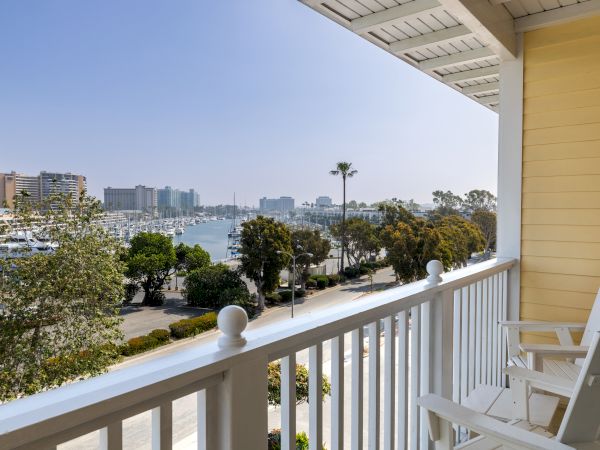 A sunny waterfront view from a balcony with white railings, palm trees, and boats along a marina, overlooking calm water and distant buildings.