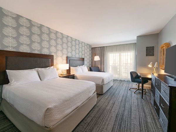 The image shows a hotel room with two beds, a desk, a chair, a TV, and lamps, featuring patterned wallpaper and a window with curtains.