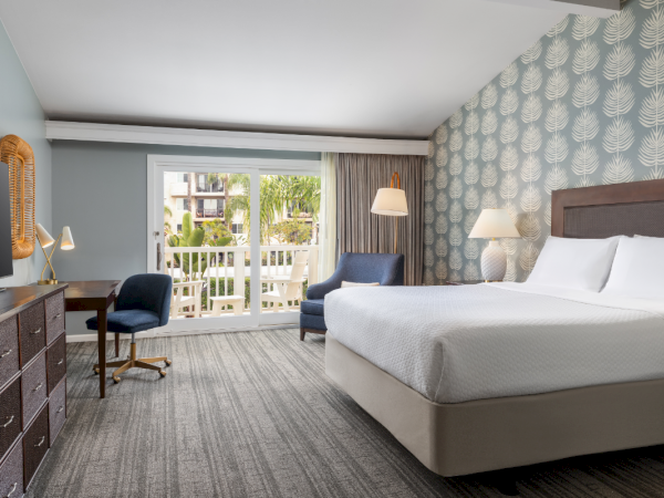 A modern hotel room with two queen beds, a desk and chair, a dresser with a mirror, and a balcony view of palm trees beyond sheer curtains.