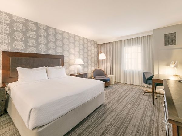 A modern hotel room with a bed, two chairs, lamps, a desk, and a window with curtains, featuring elegant wallpaper and carpet flooring.