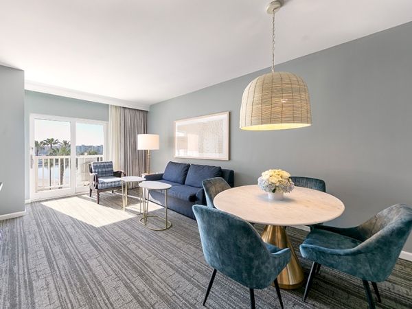 A modern living area features a blue sofa, round dining table with chairs, pendant light, and access to a balcony with natural light.