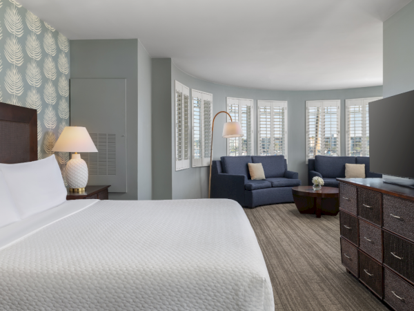 A spacious hotel room with a king bed, nightstands, a curved seating area by bay windows, blue sofas, a dresser, and a flat-screen TV, all in calming tones.