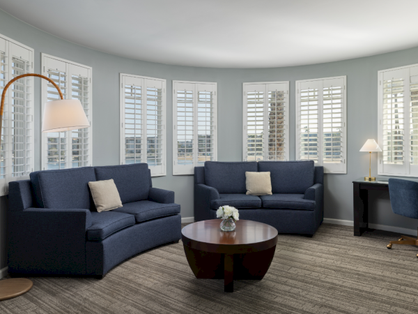 A bright living room with blue sofas, white shutters, a wooden coffee table, a tall arc floor lamp, and a small desk in the corner, cozy and airy.