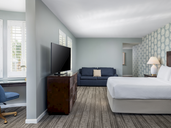 A bright hotel suite with two beds, a sofa, a TV on a dresser, a desk by windows, and soft patterned wallpaper along the far wall.