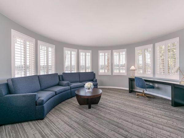 A curved room with a blue sofa, round table, and desk with a chair and lamp by shuttered windows.