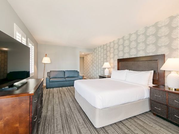 A hotel room with a bed, two nightstands with lamps, a sofa, a dresser with a TV, and patterned wallpaper.