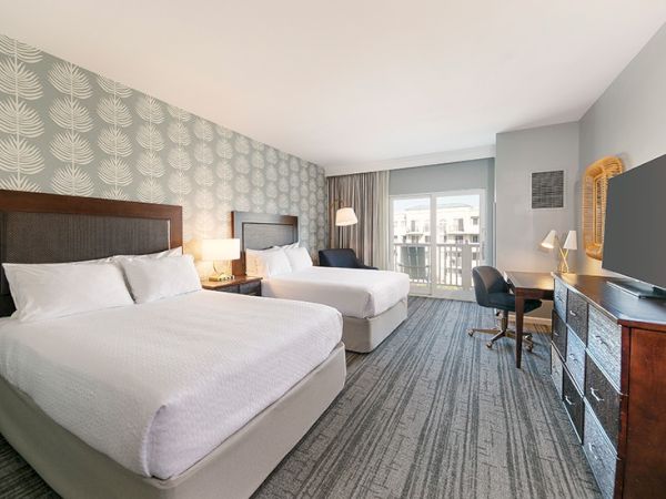 The image shows a hotel room with two beds, a desk, a TV, and a sliding door leading to a balcony, featuring modern decor.