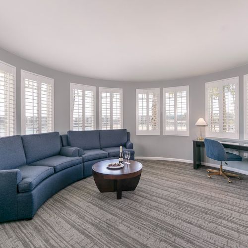 The image shows a room with a round layout, featuring a blue sectional sofa, a coffee table, a desk, and windows with shutters.