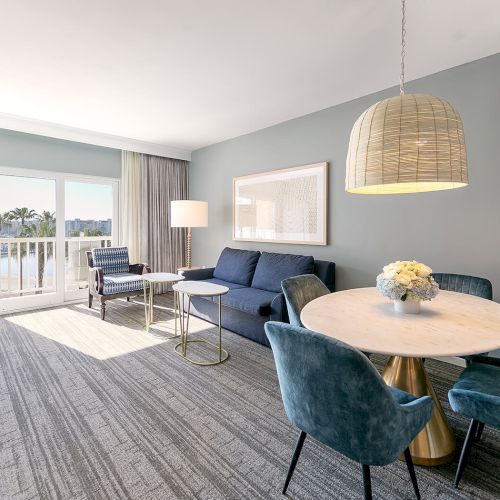 A bright living room with a sofa, dining table, chairs, and a large window leading to a balcony, featuring modern decor.