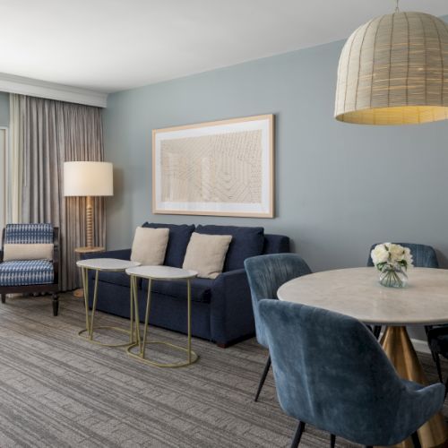 A modern hotel living area with blue walls, a sofa and chairs, a round marble table, a wicker pendant light, a framed art piece, and a balcony door.