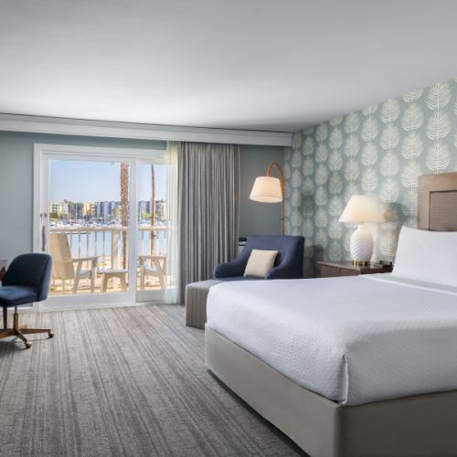 A modern hotel room with two large beds, a desk, a chair, and a balcony view of the city, calm lighting and patterned wallpaper.