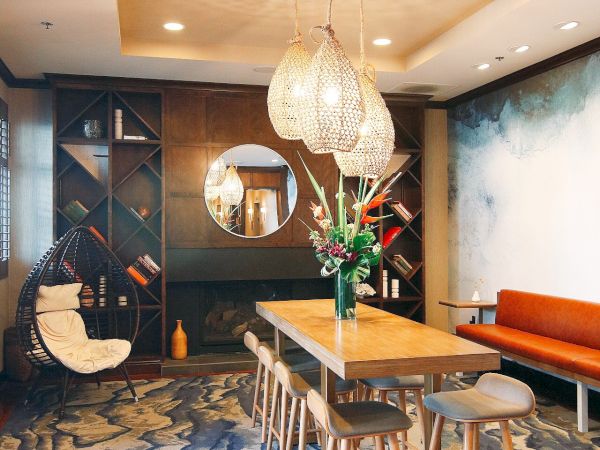 A stylish dining room with a wooden table, stools, pendant lights, abstract carpet, and wall art, with a mirror and bookshelf.
