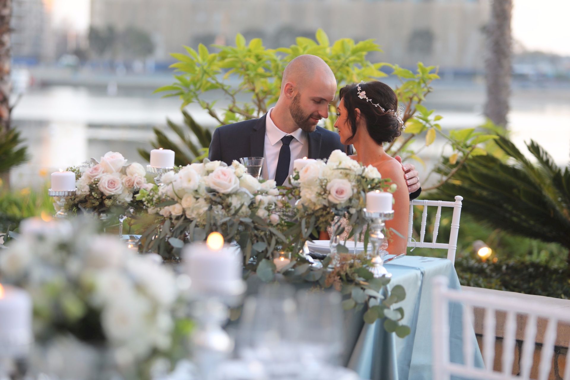 A bride and groom share a quiet moment at an outdoor wedding dinner, surrounded by roses, greenery, and candlelight.