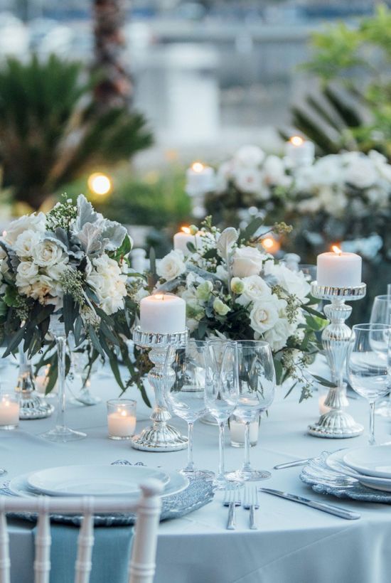 Elegant outdoor banquet setup with round tables, white blooms, candles, and glassware on pale blue linens, amid greenery and white chairs.