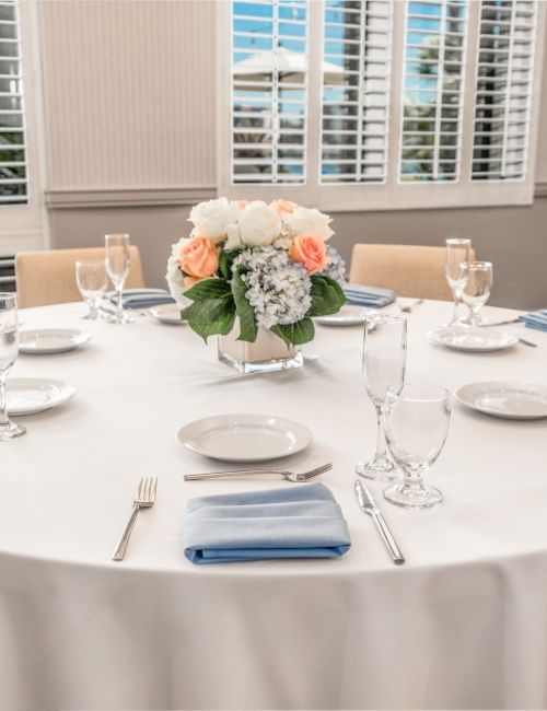 A round table is set for a formal meal with white linens, glassware, silverware, and a floral centerpiece in a bright, elegant dining room.