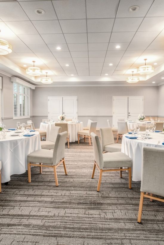 A bright, airy banquet room with round tables draped in white linens, gray upholstered chairs, blue place settings, and soft overhead lighting.