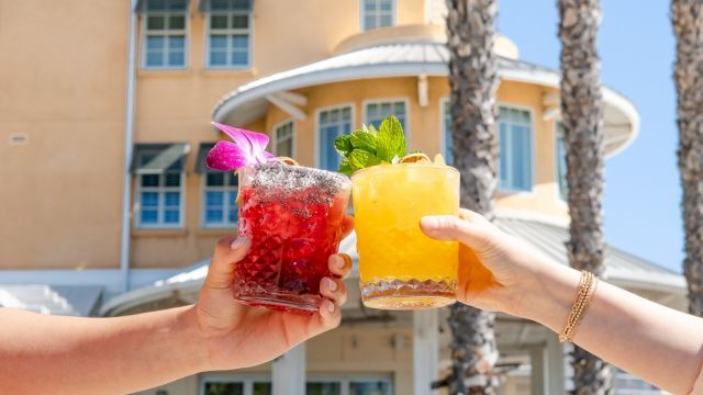 Two hands clink colorful cocktails in front of a sunny hotel, with garnishes and a bright, tropical vibe. End with a period.