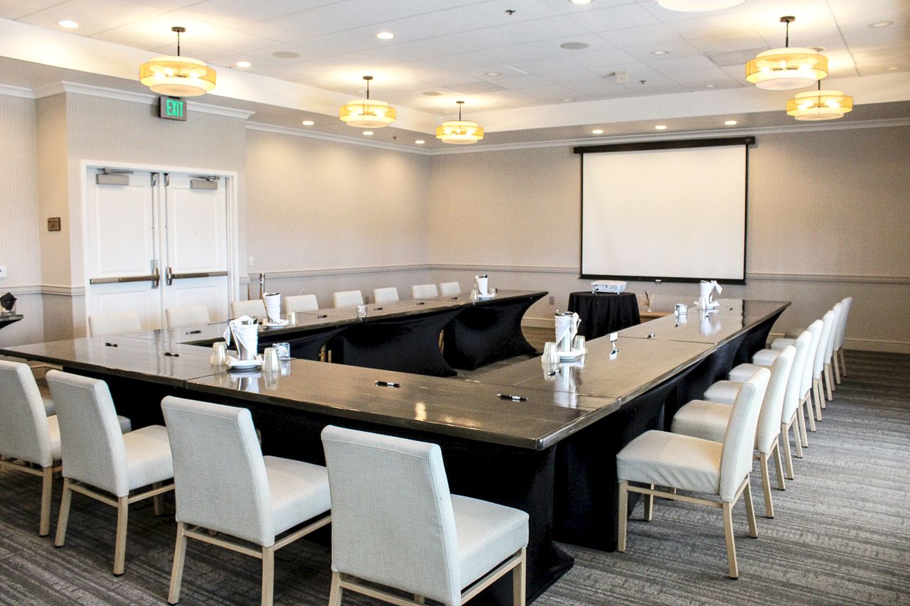 A modern conference room with a large boardroom table, cushioned chairs, and a projector screen under ceiling lights.