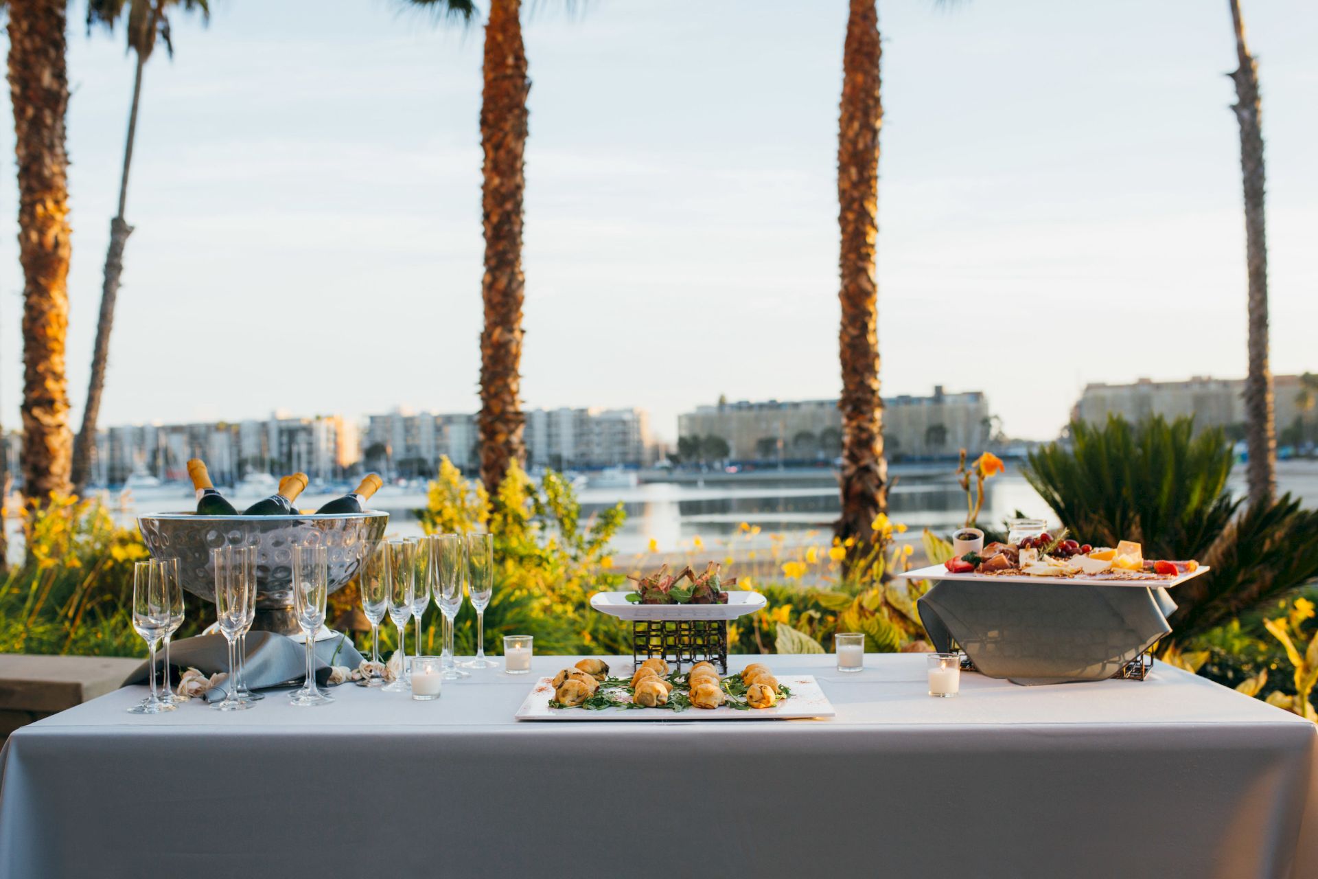 Outdoor dining setup with a tub of champagne, sparkling flutes, and buffet platters on a table among palm trees overlooking a city skyline in golden light.