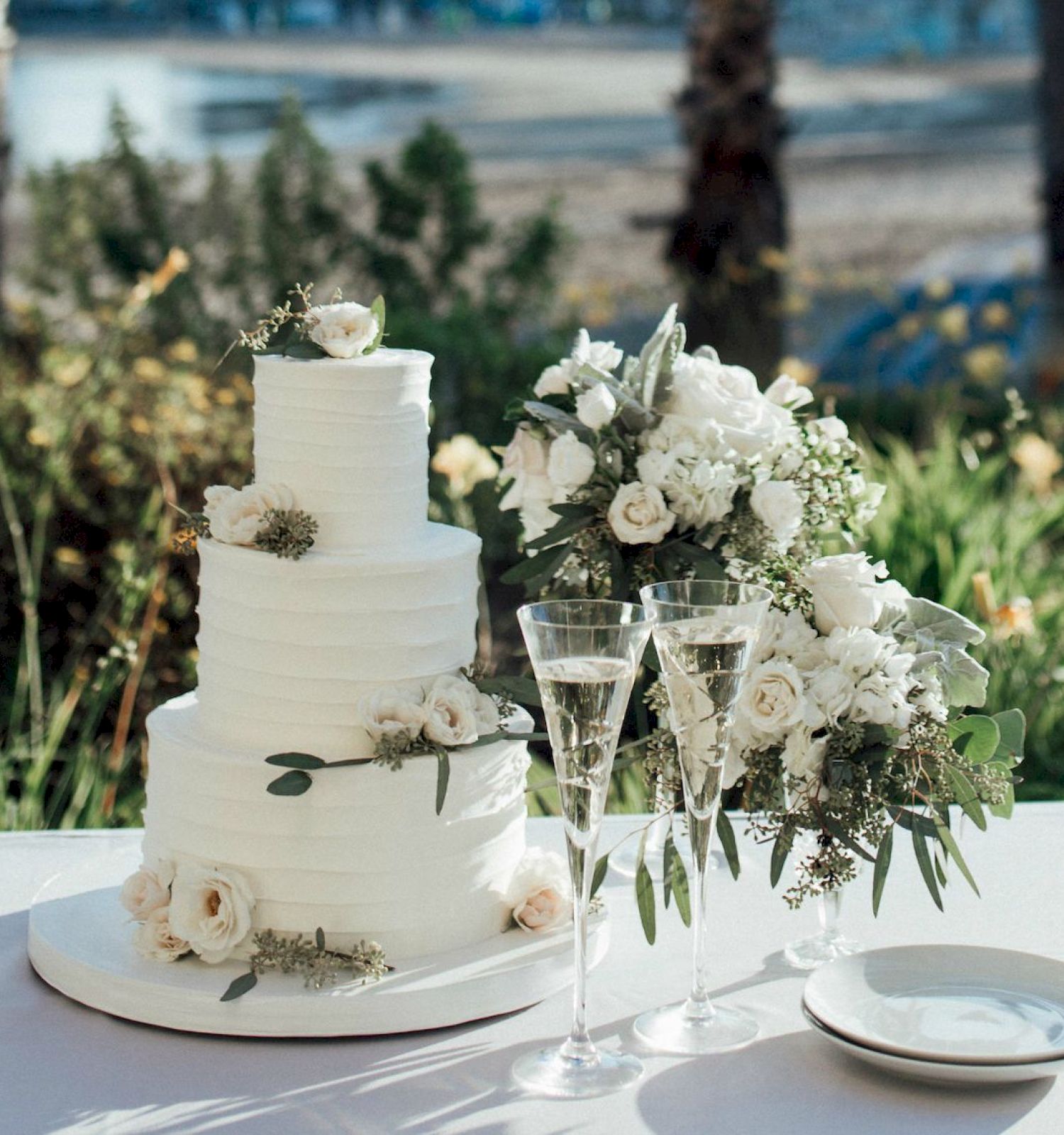 A wedding cake on a table with floral decorations and a beach backdrop with palm trees and a distant building. Ending punctuation.
