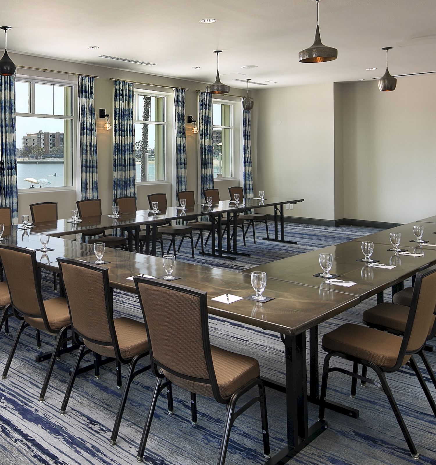 A long U-shaped conference table setup in a bright meeting room with windows, chairs, and place settings, overlooking water.