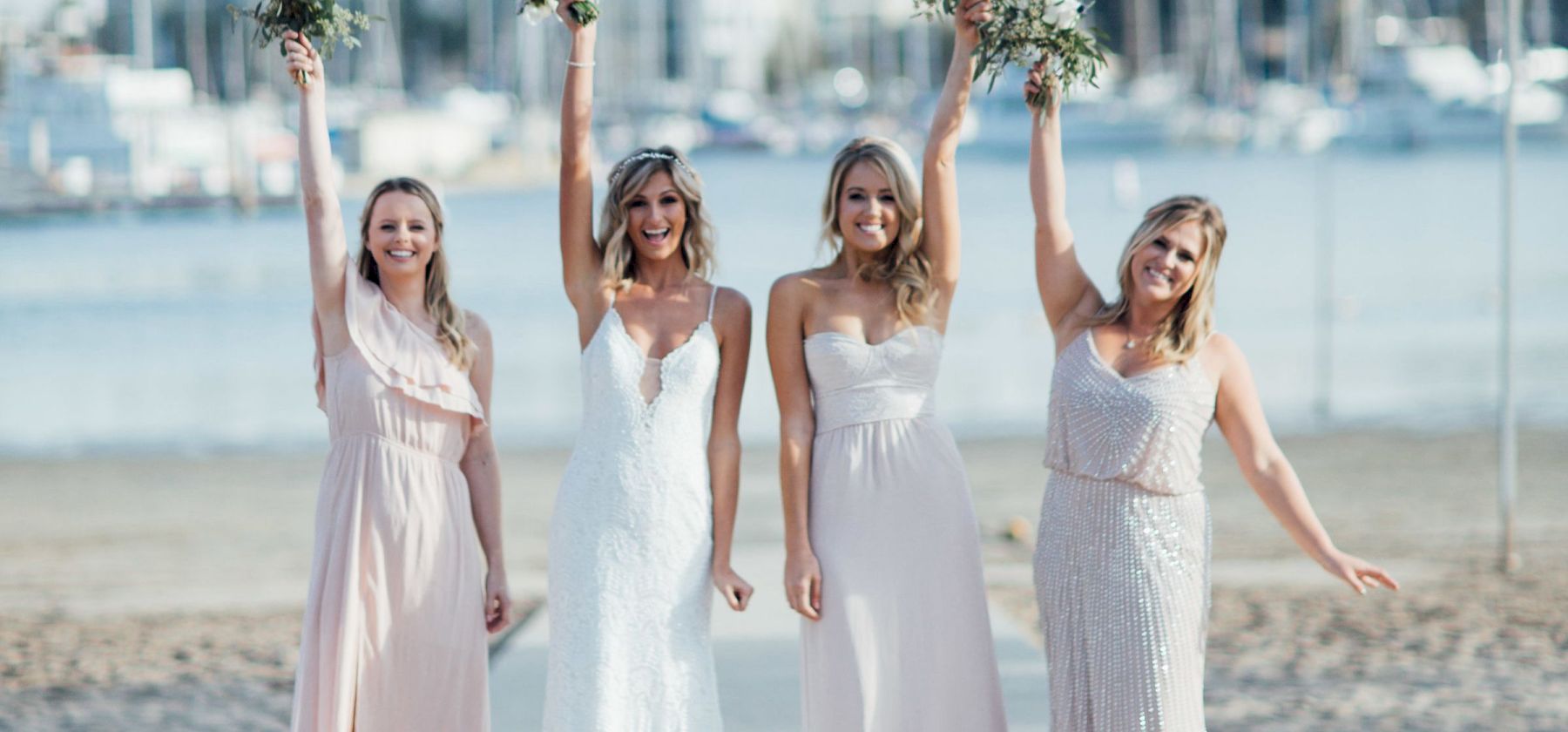 Four women in white dresses stand on a beach, cheering and holding bouquets in the background of the waterfront and city skyline.