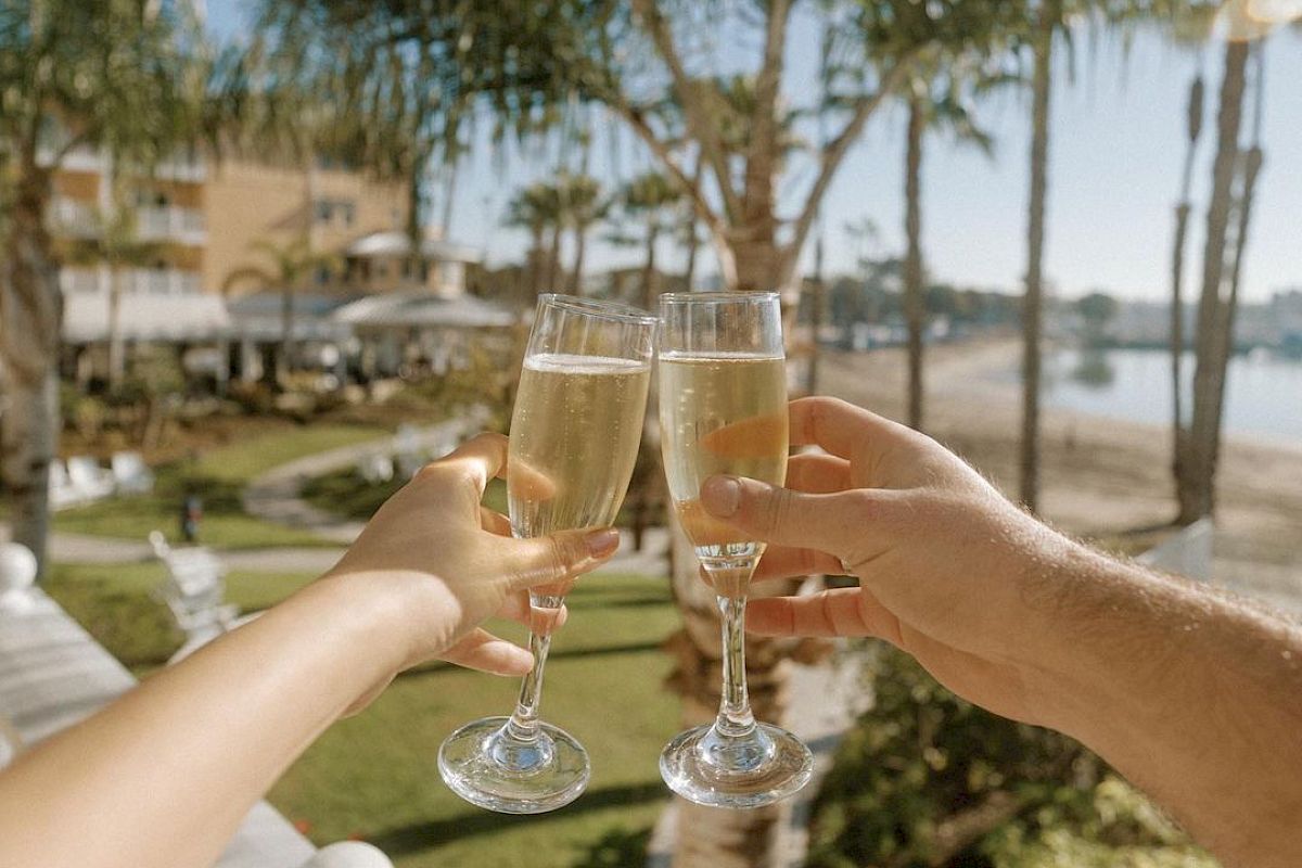 A toast on a sunny balcony by the water, palm trees and champagne glasses clinking as friends celebrate together.