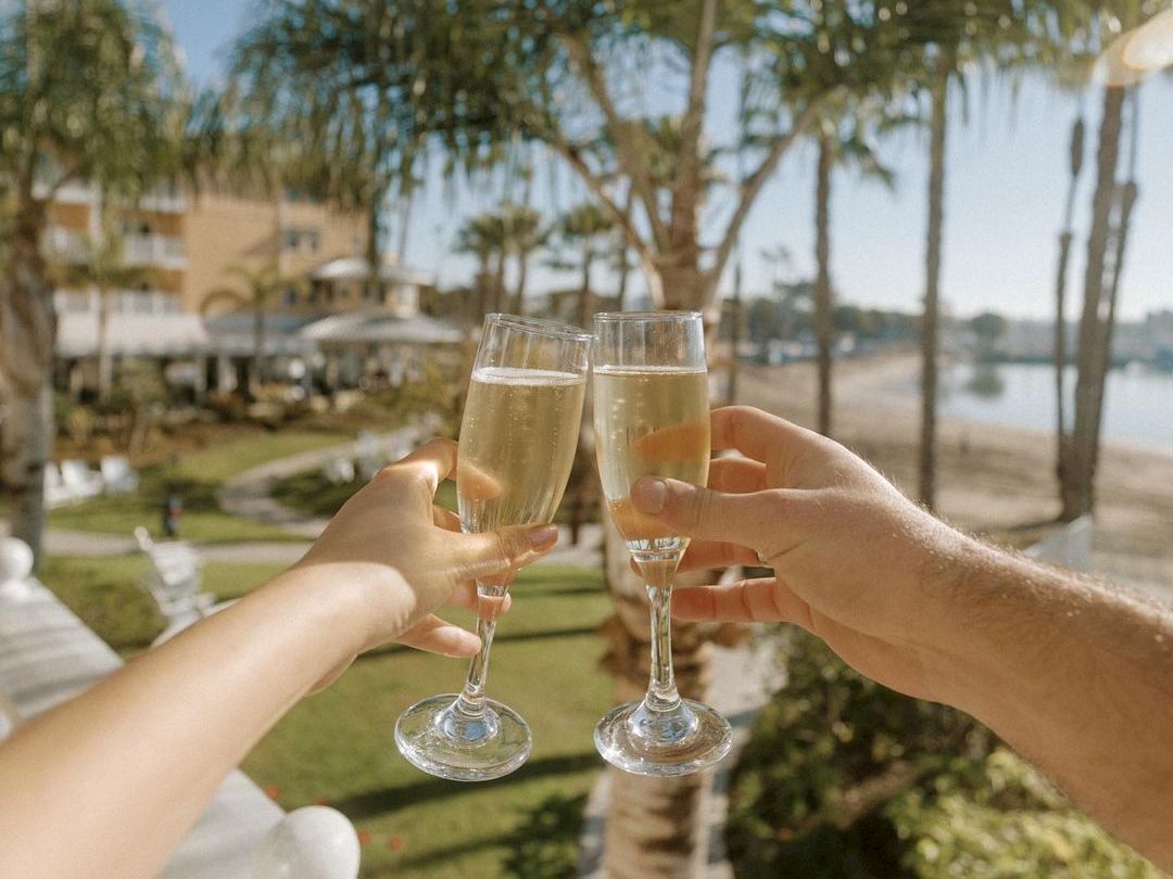 A toast on a sunny balcony by the water, palm trees and champagne glasses clinking as friends celebrate together.