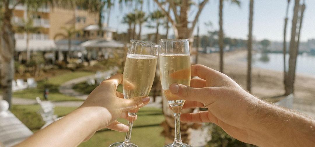 A toast on a sunny balcony by the water, palm trees and champagne glasses clinking as friends celebrate together.