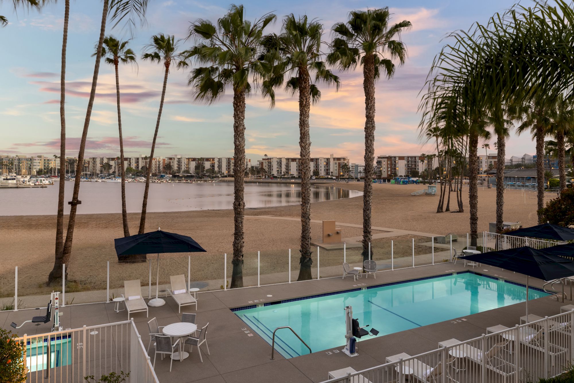 A beachside pool with white fencing, palm trees, empty chairs, and a calm shoreline in the background at sunset.