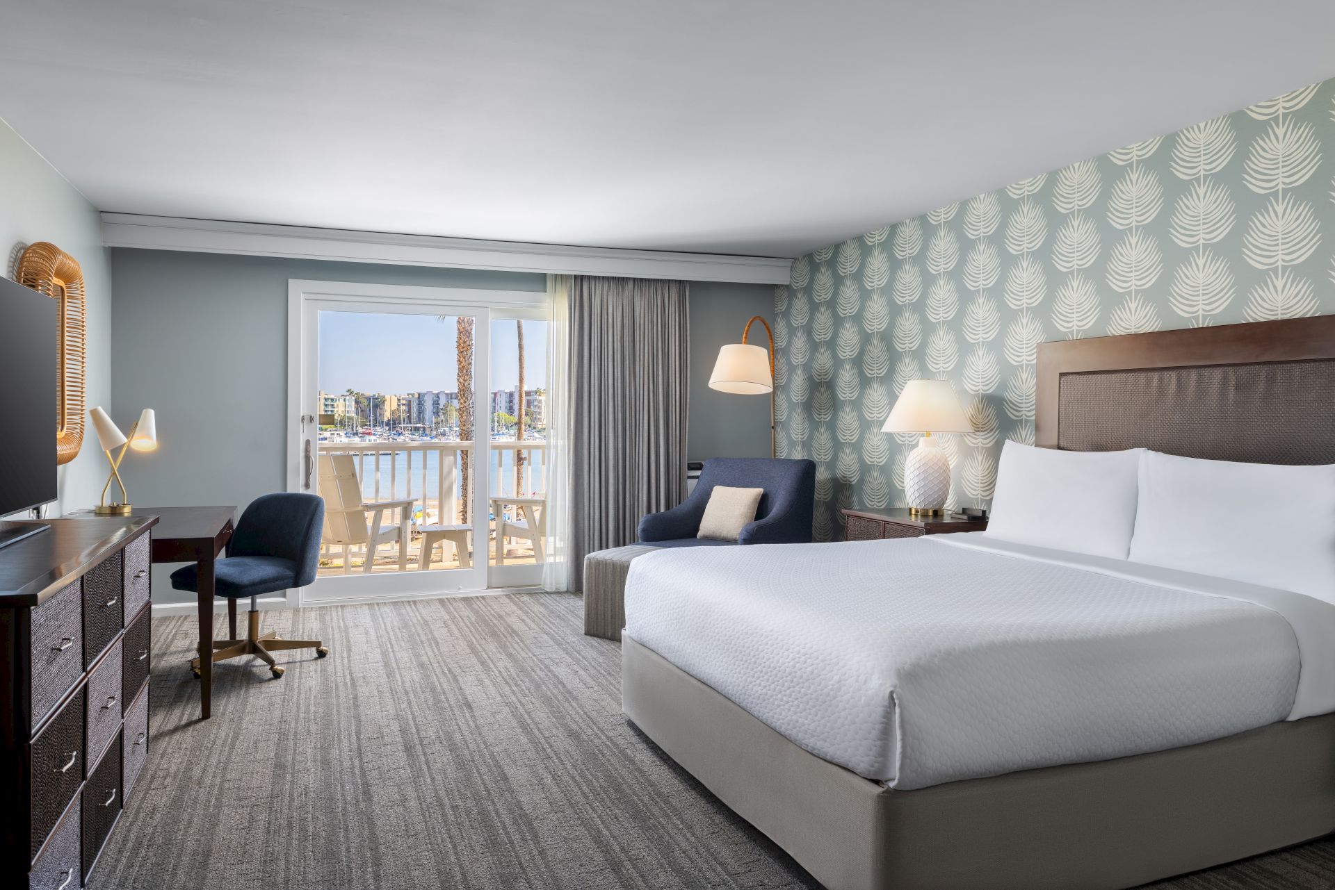 A modern hotel room with two queen beds, a desk and chair, a seating area, patterned wallpaper, and a balcony with city views.