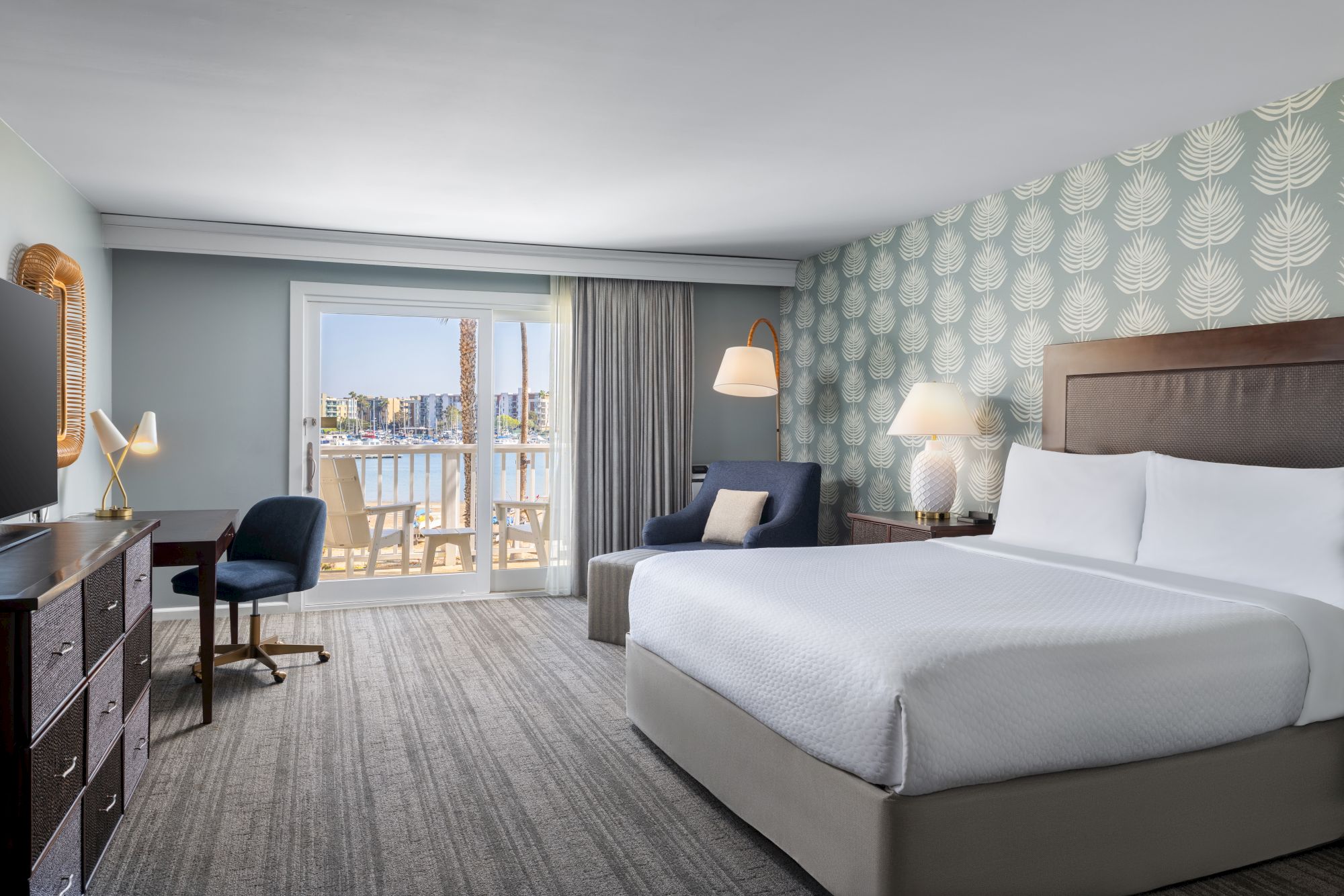 A modern hotel room with two queen beds, a desk and chair, a seating area, patterned wallpaper, and a balcony with city views.