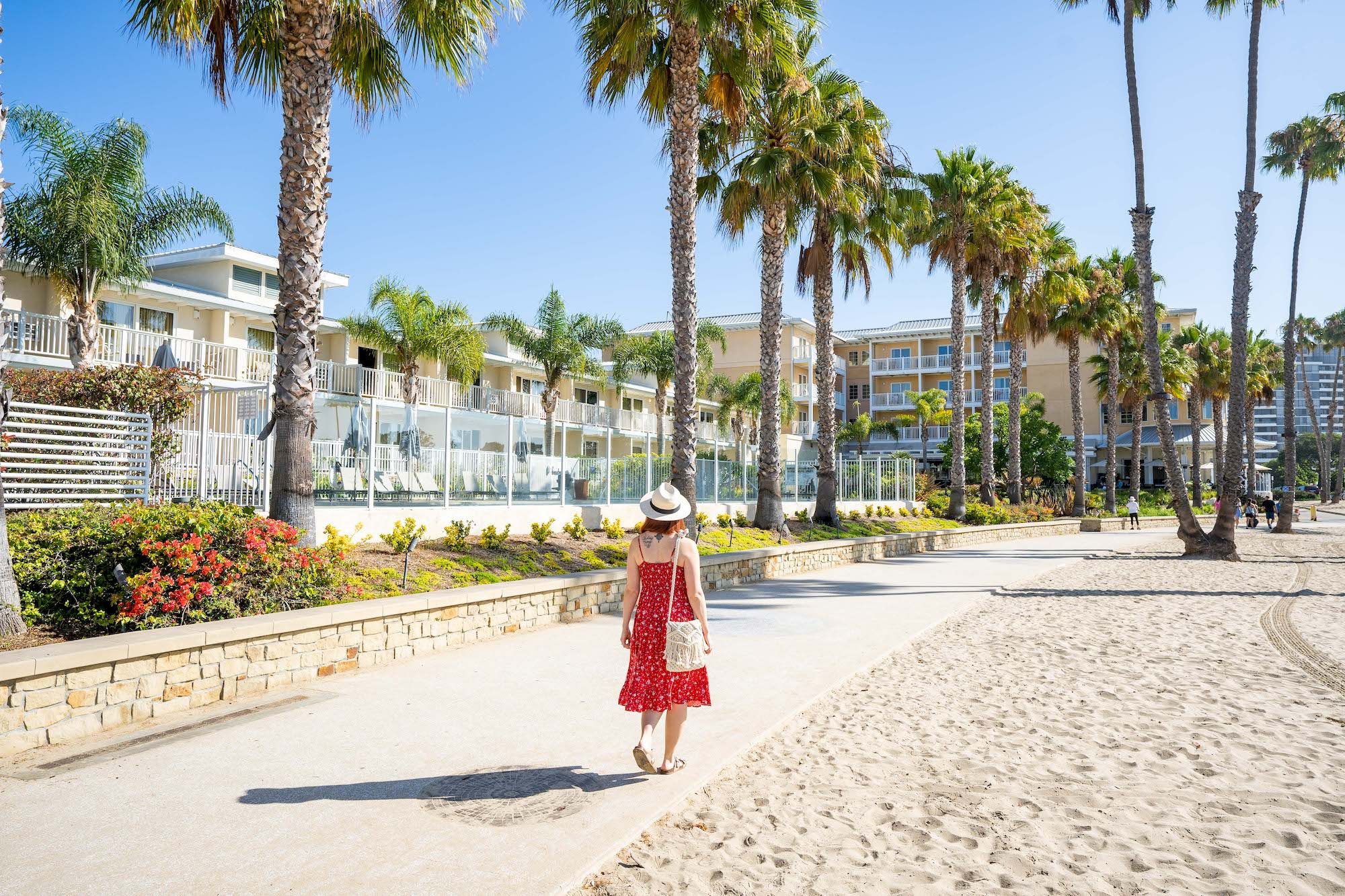 A woman in a red dress and hat walks along a sunny beach promenade with palm trees, water, and resort buildings in the background.