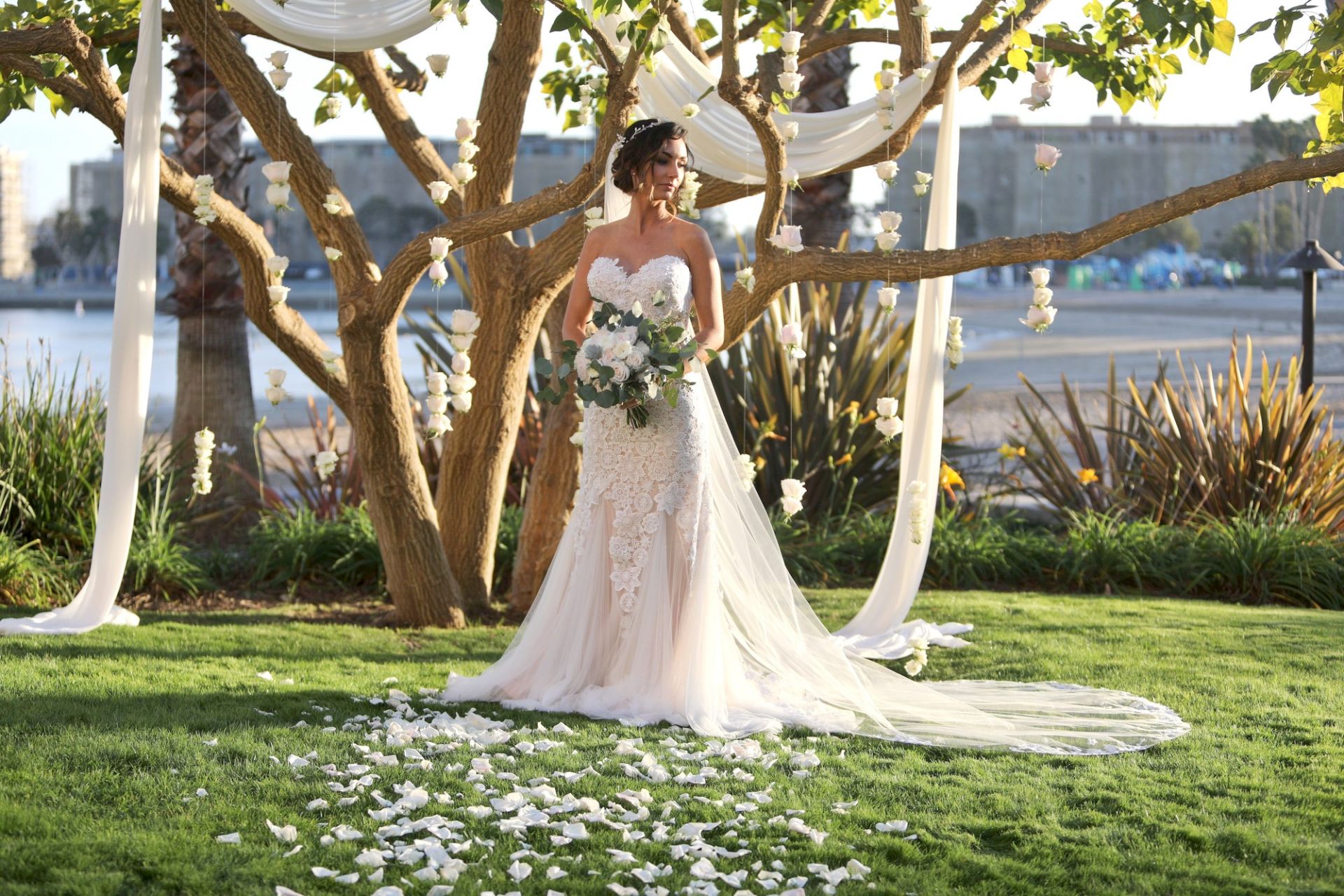 A bride in a white lace gown stands beneath hanging roses and sheer drapes in an outdoor garden wedding ceremony.